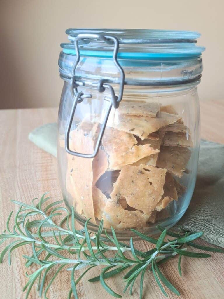 Rosemary olive oil sourdough crackers stored in a clear glass air tight jar.