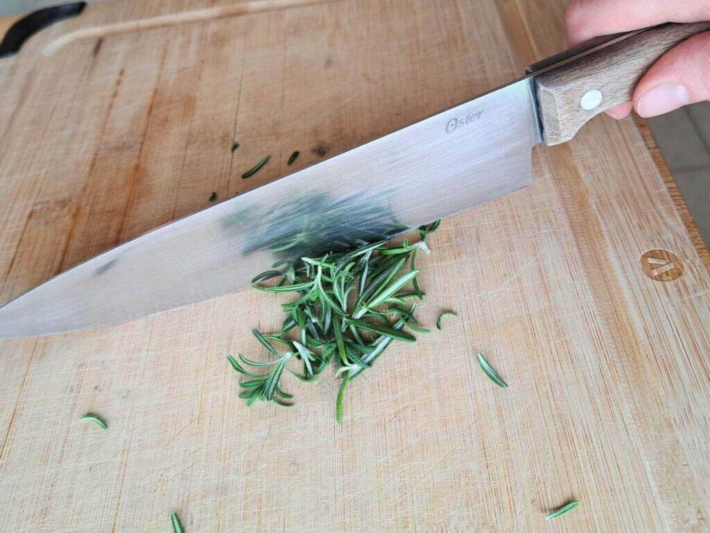 Finely chopping up fresh rosemary leaves on a wooden cutting board.