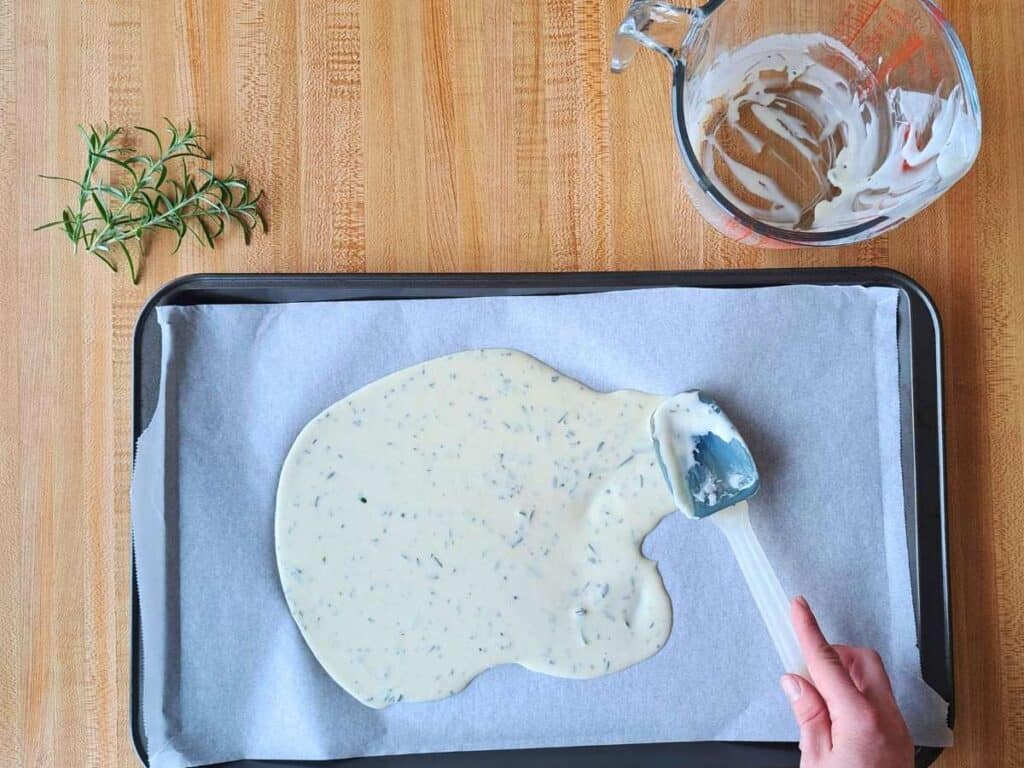 Spreading the discard cracker mix out on a parchment lined baking sheet next to an empty glass measuring cup.