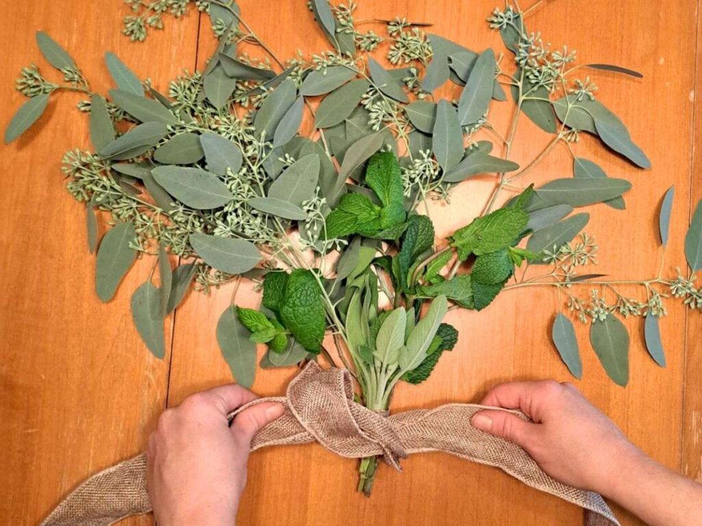 Tying a burlap bow around a bunch of fresh cut herbs that are laying on a wooden table.