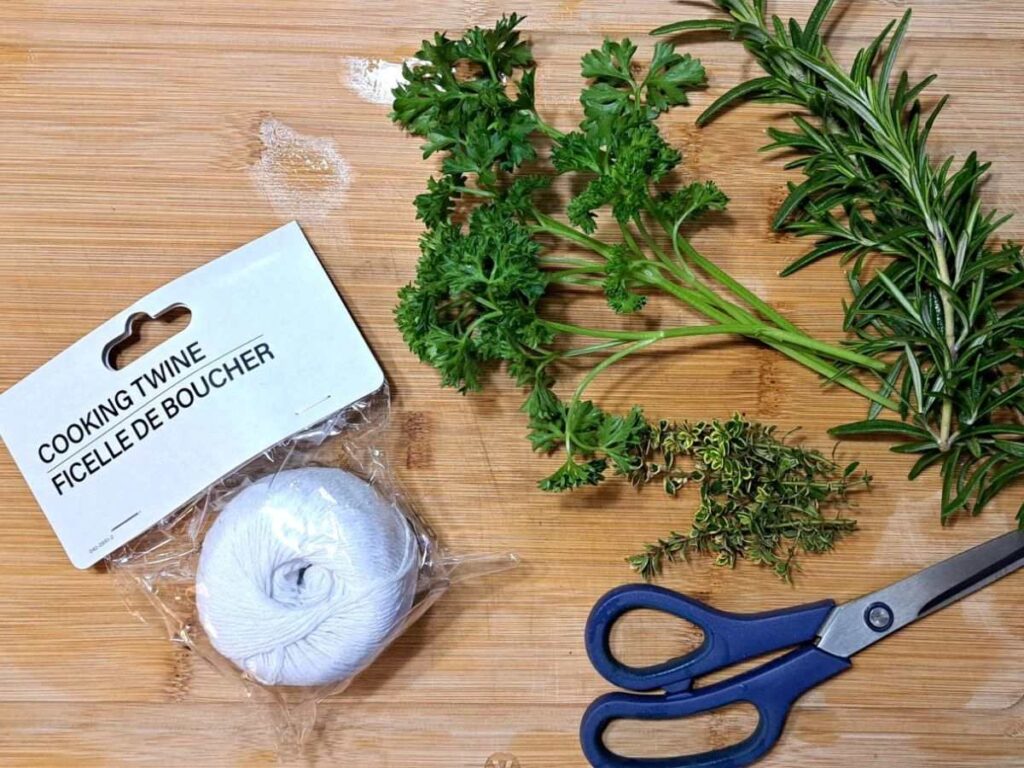 Supplies gathered on a cutting board including cooking twine, scissors, and freshly cut herbs.