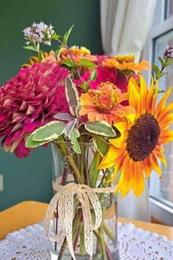 A mix of herbs and summer flowers in a clear glass vase sitting on a wooden table.