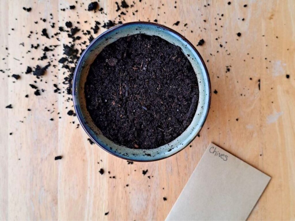 A pot with dirt in it on a wooden table with a packet of chive seeds beside it.