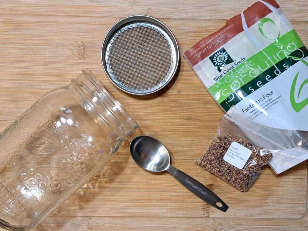 Supplies to grow sprouted herbs in a jar all laid out on a wooden cutting board.