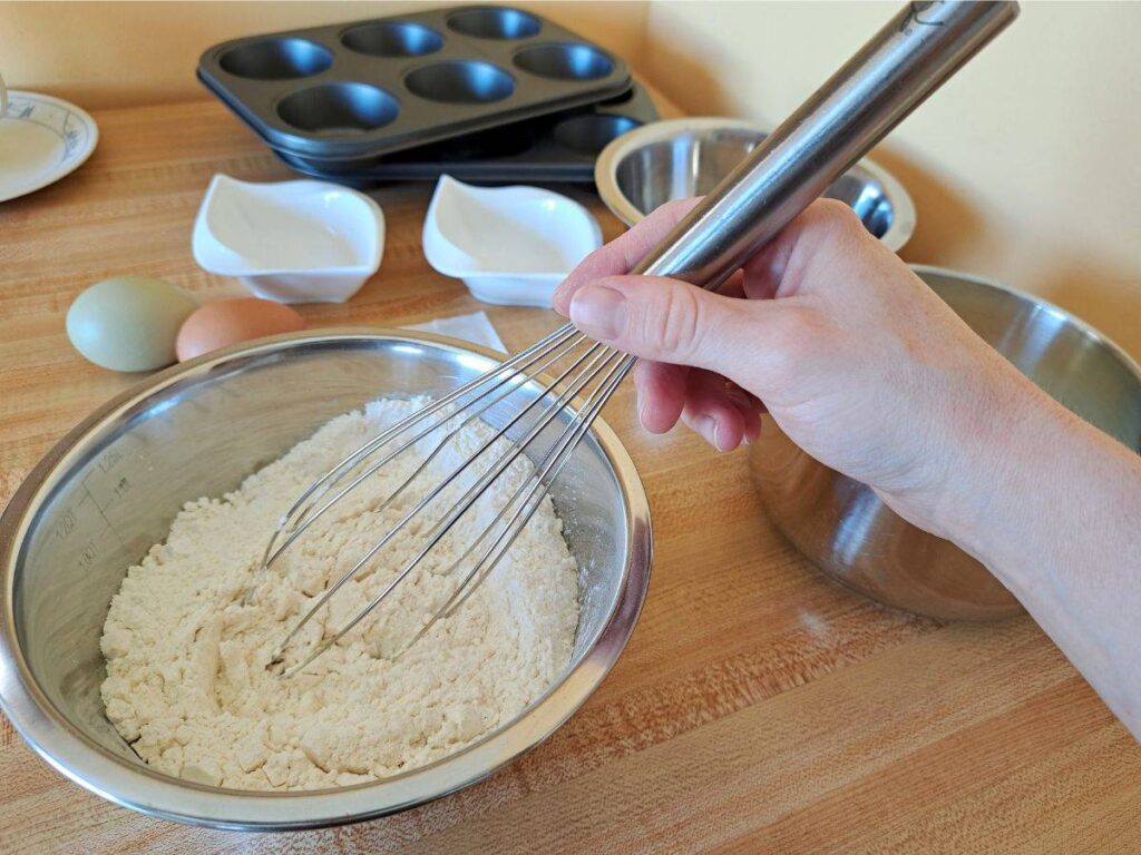 Someone mixing flour, and other dry ingredients in a metal bowl.