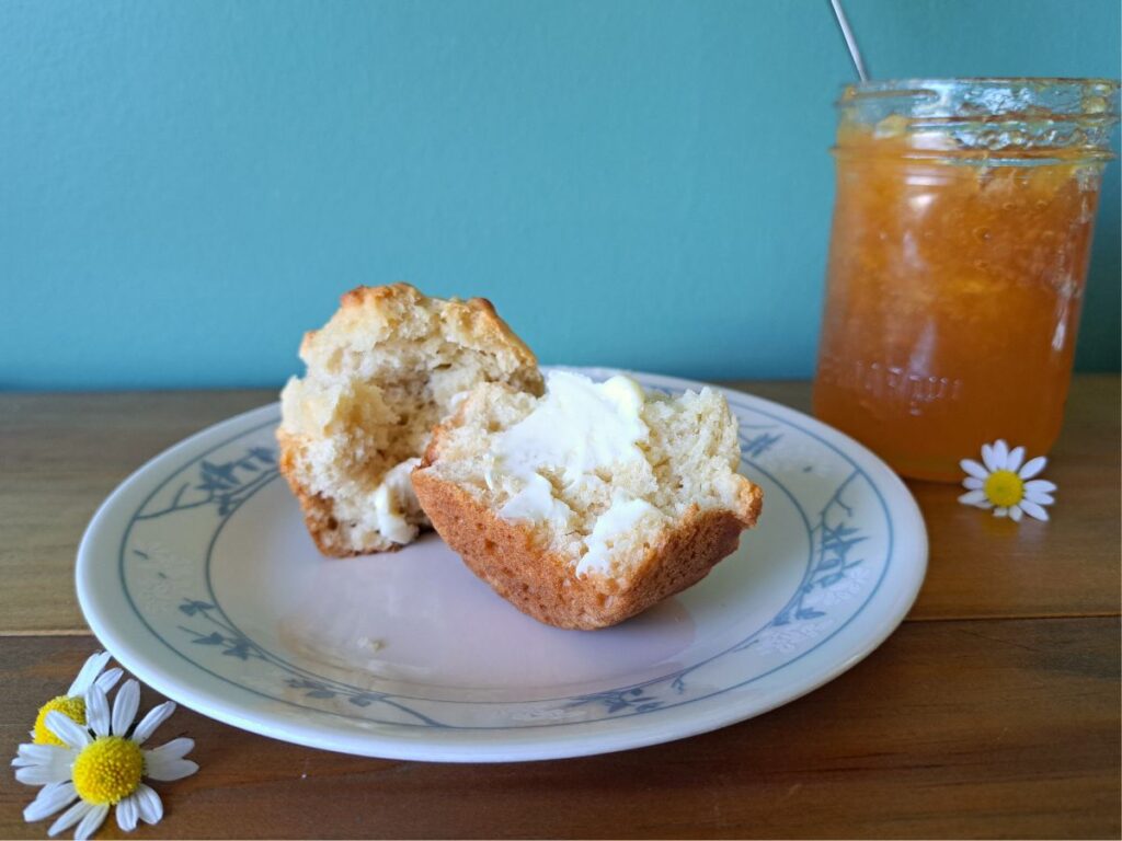 A chamomile honey sourdough muffin cut open on a plate with butter on it and peach jam in the background.