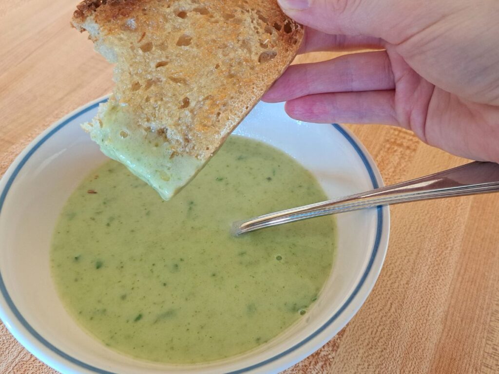 A close up of a piece of toast dipped into cream of potato and parsley soup.