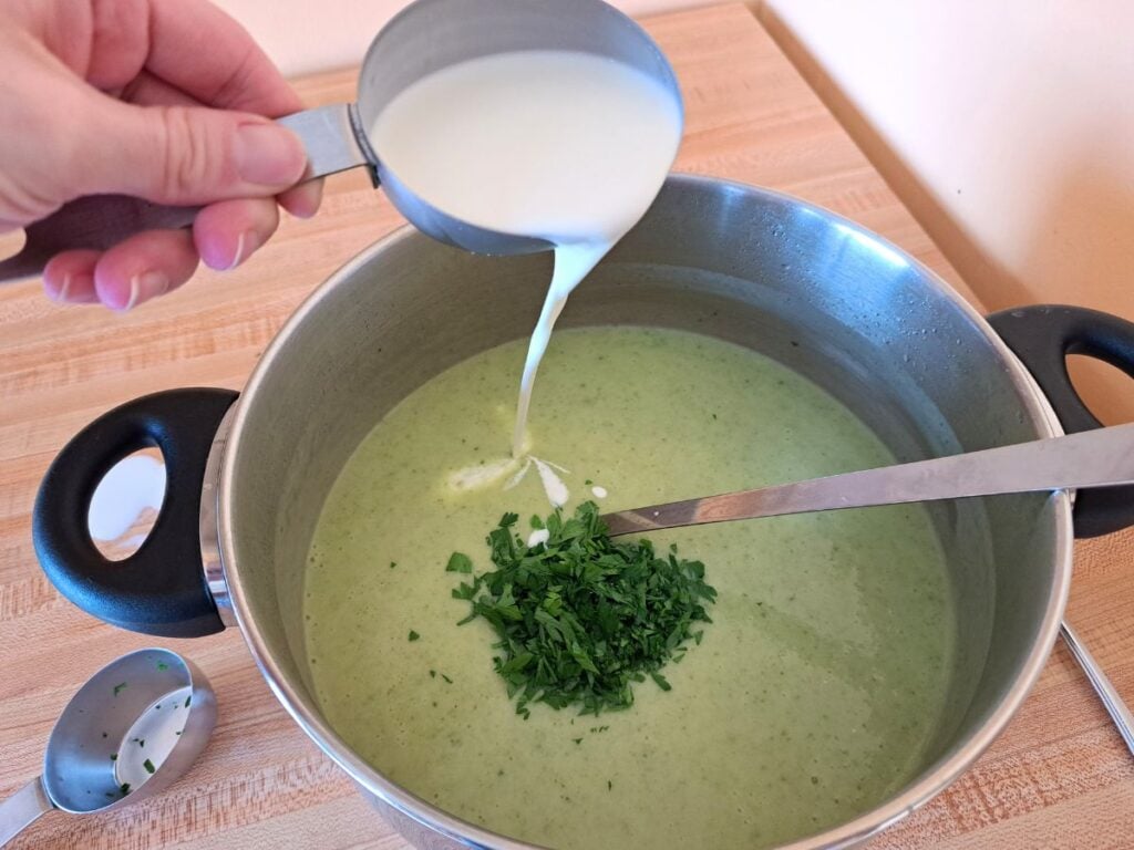 A close up of soup in a pot with parsley in it. Cream is being poured in to it.