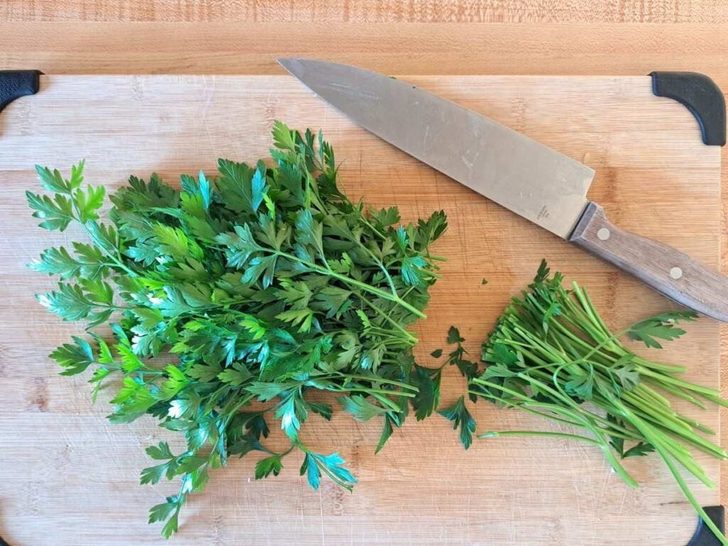A cutting board with a knife and parsley stems cut off.