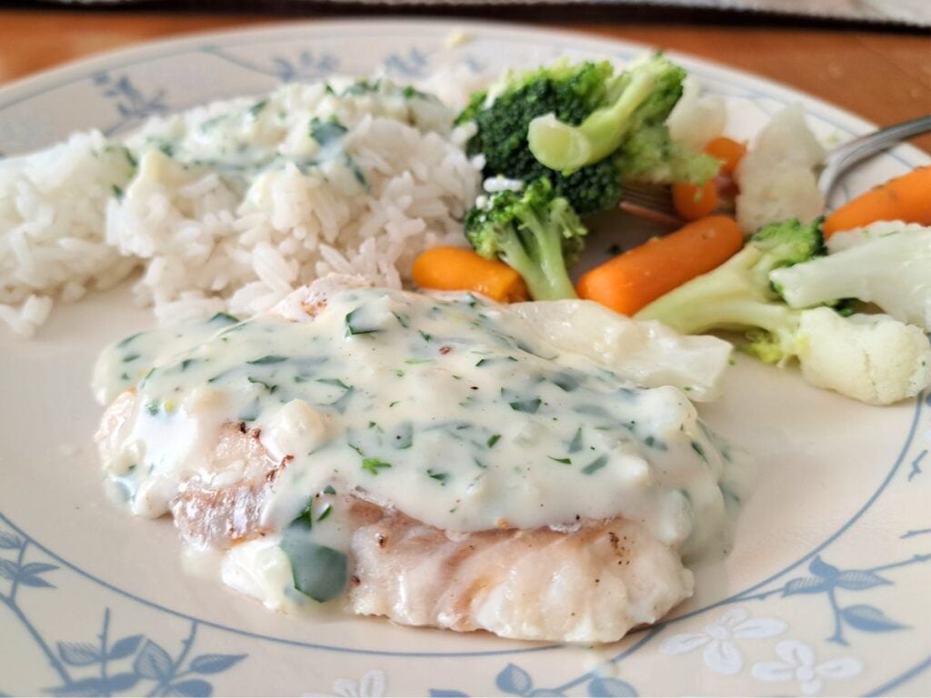 A dinner plate with white fish in parsley sauce, rice, and mixed vegetables.