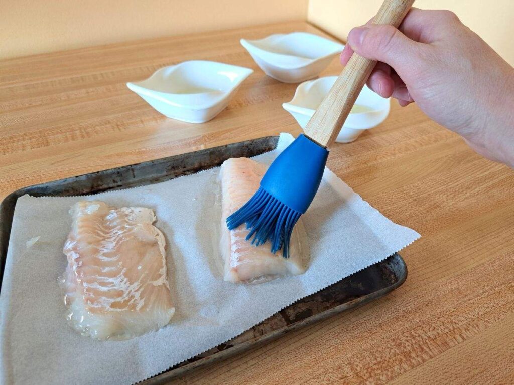 Brushing fish fillets with olive oil on a parchment lined baking sheet.