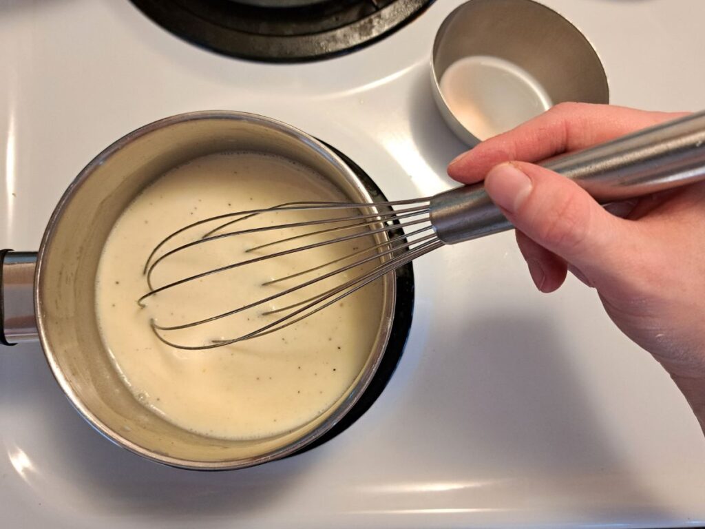 Whisking milk into the roux in a metal pot on the stove.