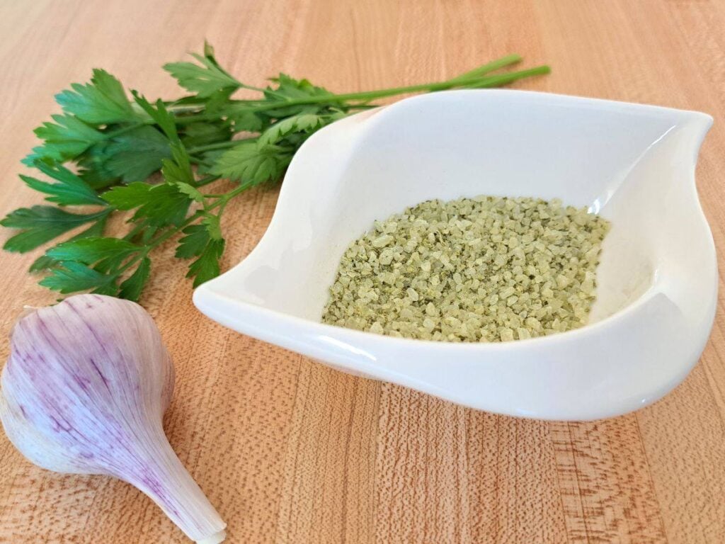 A close up of homemade garlic parsley salt in a white dish with garlic and parsley on the counter beside it.
