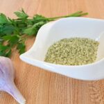 A close up of homemade garlic parsley salt in a white dish with garlic and parsley on the counter beside it.