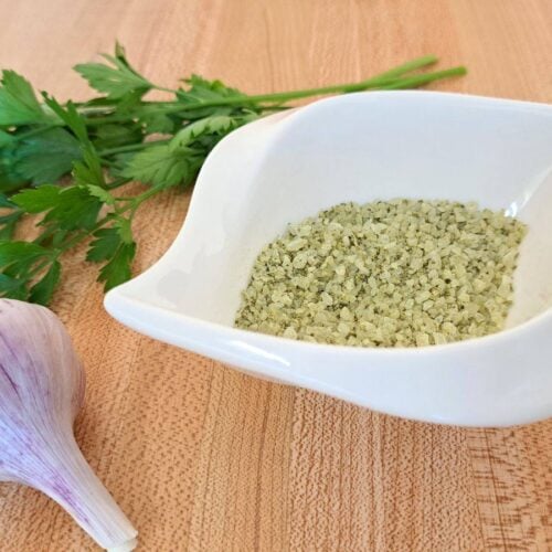 A close up of homemade garlic parsley salt in a white dish with garlic and parsley on the counter beside it.