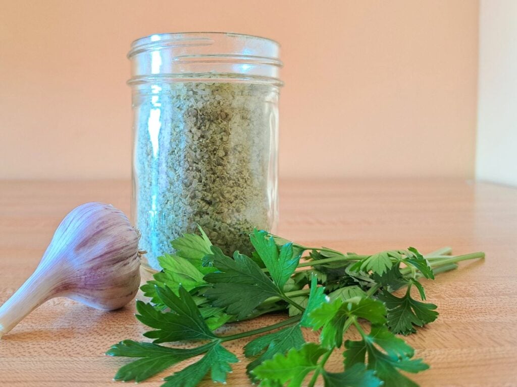 Garlic Parsley finishing salts in a jar with parsley and garlic beside it on a wooden counter.