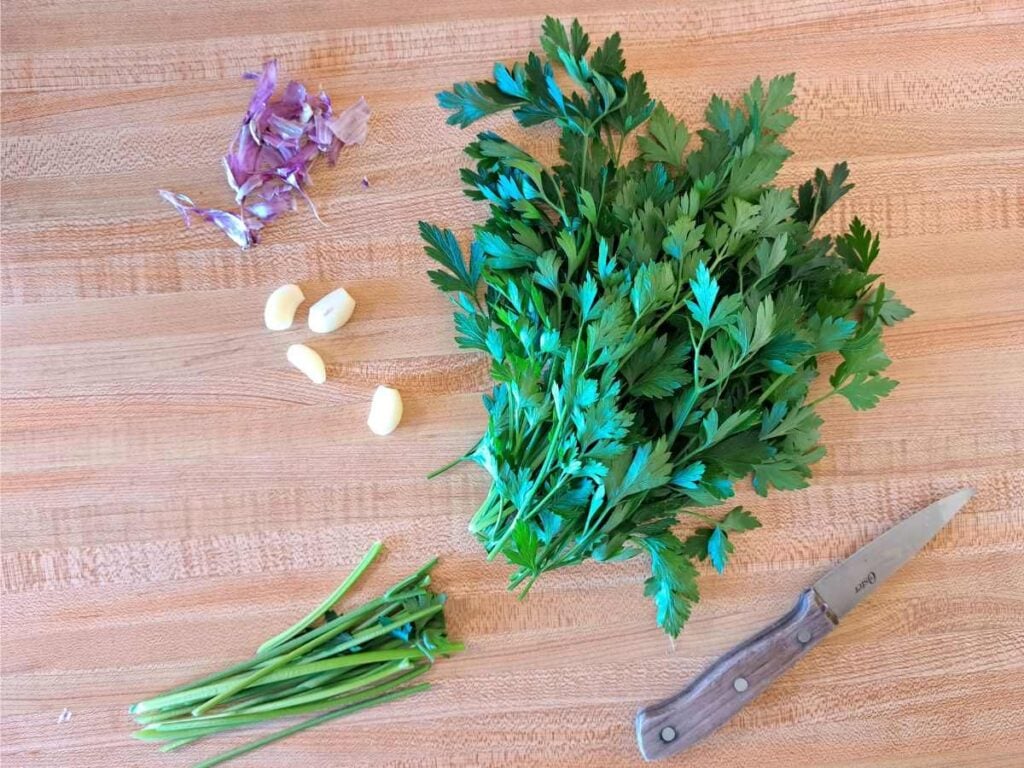 Parsley with stems cut off, and peeled garlic on a counter with a knife.