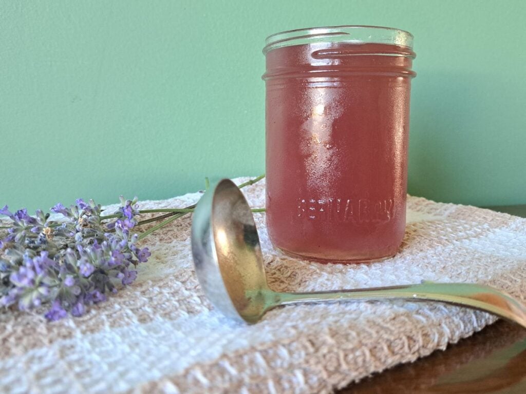 A close up of honey lavender syrup in a clear glass canning jar that is sitting on a tea towel.