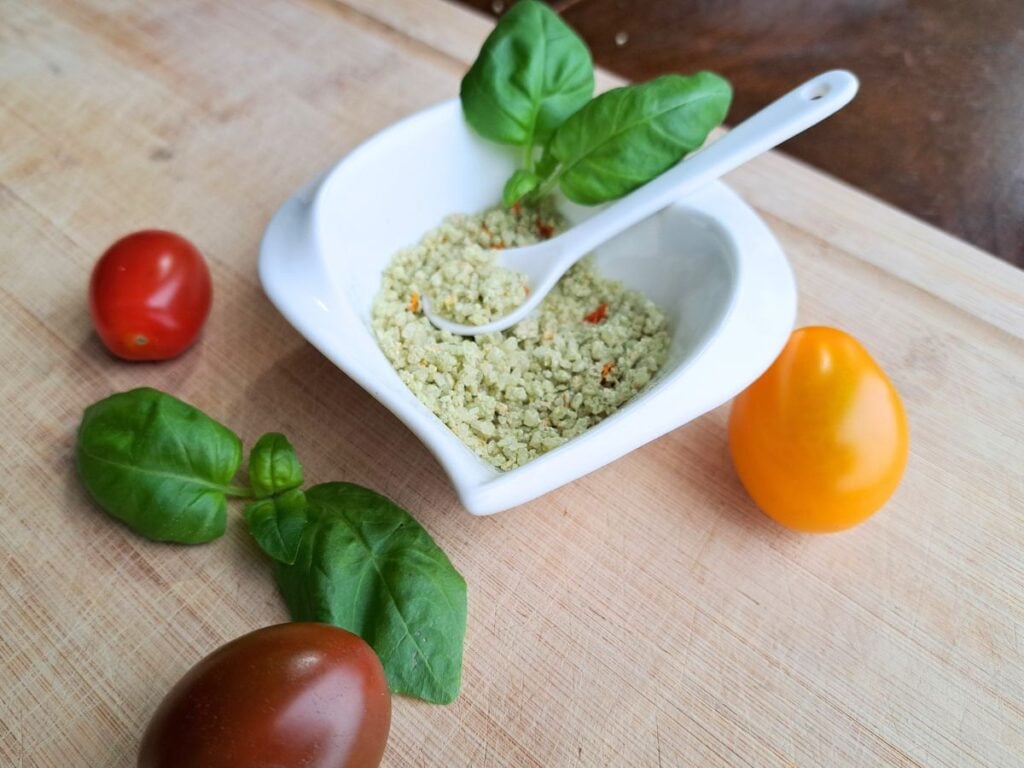 Tomato basil salt in a white dish with a spoon. Basil and tomatoes on the counter beside it.