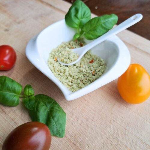 Tomato basil salt in a white dish with spoon. Basil and tomatoes on the counter beside it.