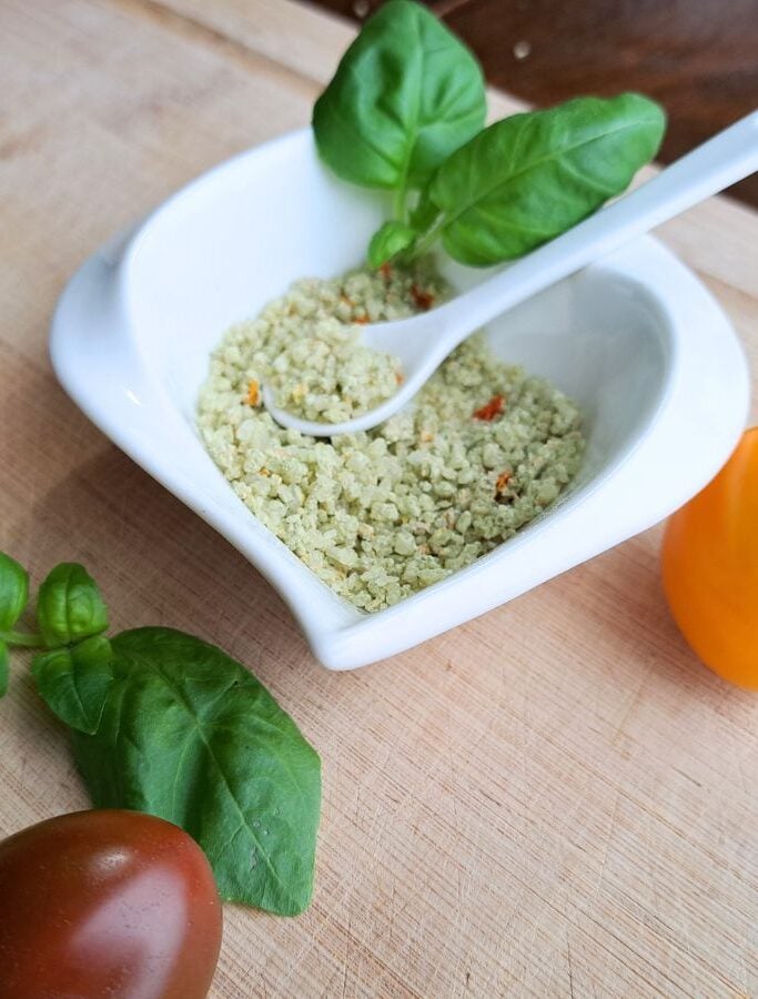 Tomato basil salt in a white dish with spoon. Basil and tomatoes on the counter beside it.