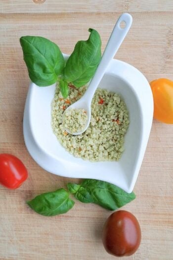 Herb sea salt in a small white dish with a spoon. There is also basil and tomato scattered around it.