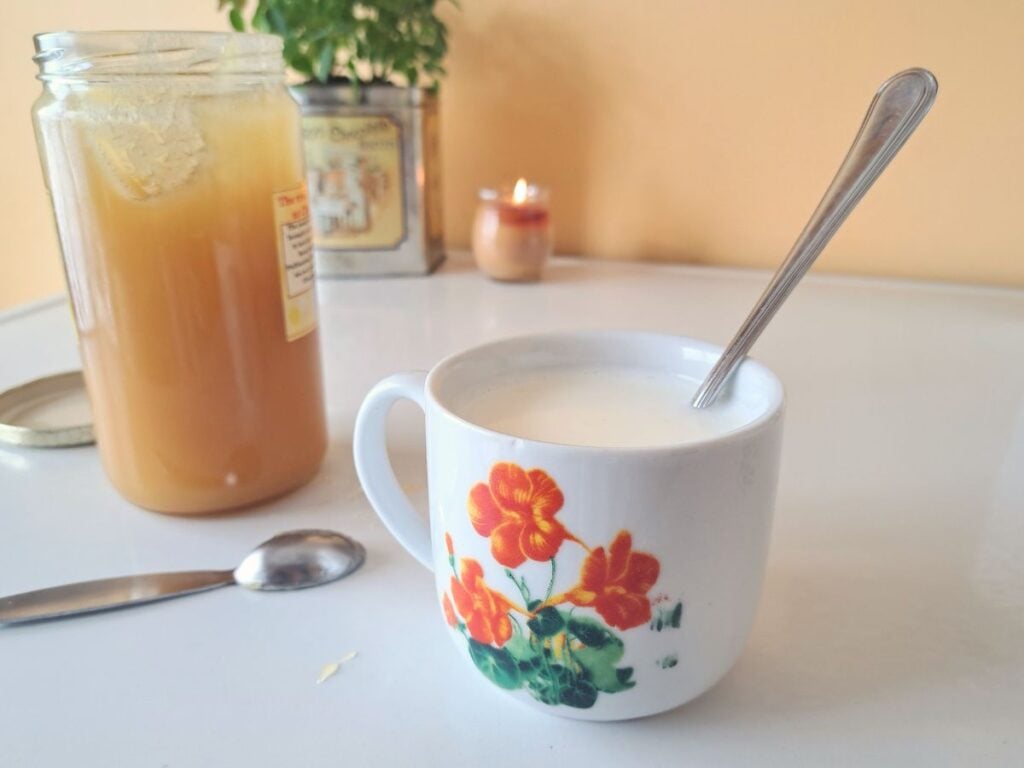 A mug with milk sitting on a counter with a spoon in it. There is a jar of honey beside it.