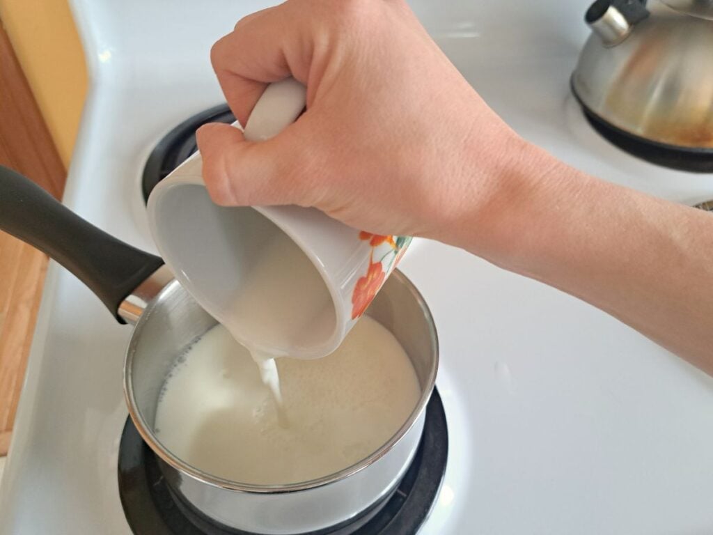Pouring milk from a mug into a saucepan on the stovetop.