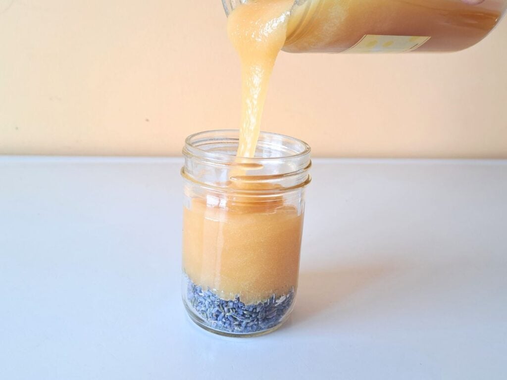 Honey being poured in to a clear glass jar with lavender buds in the bottom of it.
