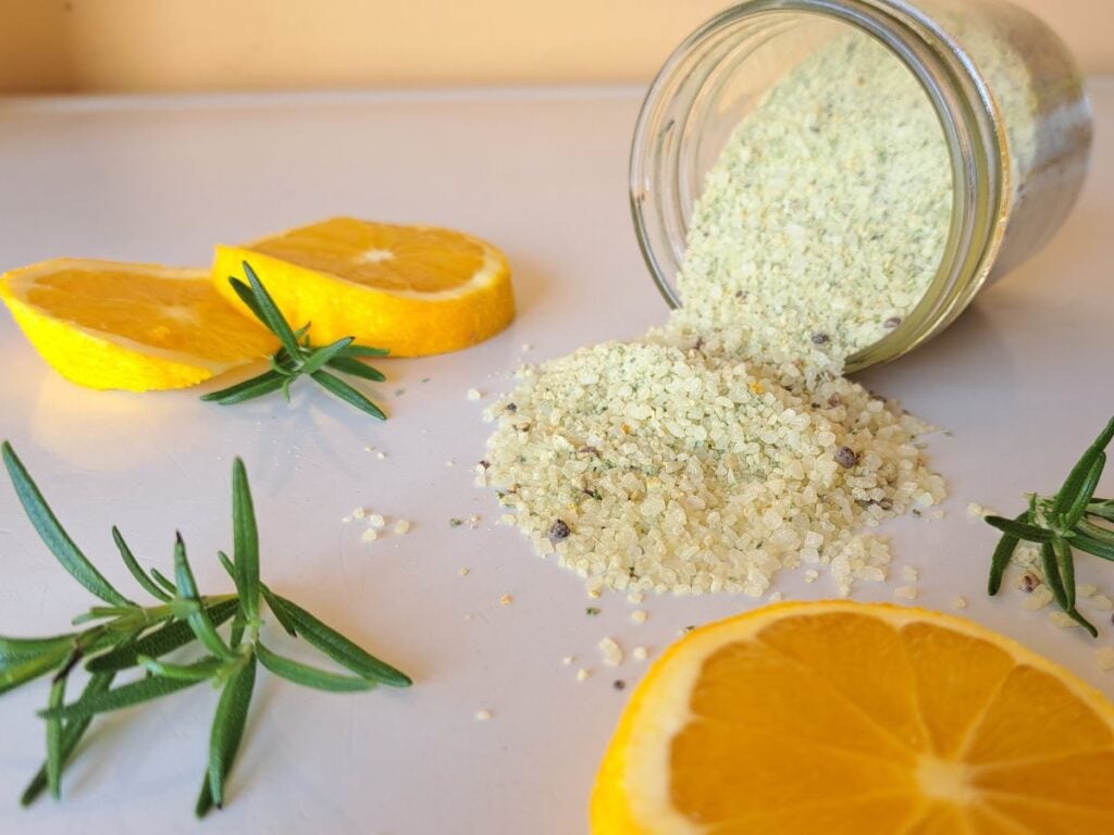Herb infused salt spilling out of a glass jar on to a counter with rosemary sprigs and orange slices beside it.