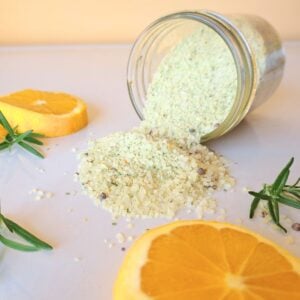 Rosemary orange salt spilling out of a container with orange slices and rosemary sprigs beside it.